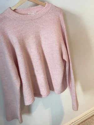 Ljusrosa stickad tröja - Mysig ljusrosa stickad tröja med rund halsringning och breda ribbade muddar vid ärmslut och nederkant. Tröjan har en avslappnad passform och är perfekt för kyliga dagar. Materialet ser ut att vara en mjuk ull- eller akrylblandning.
