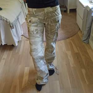 Beige camouflagemönstrade cargo byxor från Broadway. Low waist och bootcut. Justerbara snören vid bensluten. Minimal fläck vid gylfen, samma vid bensluten, inte provat ta bort. Snygga broderade detaljer fram och bak saknas några dekorativa stenar, märks knappt. Storlek M, jag på bilden brukar bära XS och är 160, de är stora i midjan och lite för långa. Passar någon som bär S-M och är ca 165 cm+. Mått: innerbenslängd: 80 cm midja omkrets: 85 cm grenhöjd: 20 cm 