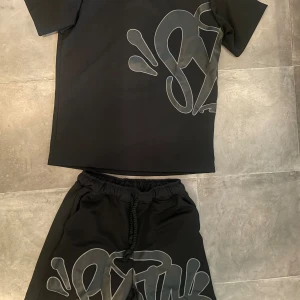 Syna world - Detta är ett trendigt svart set från Syna World, bestående av en kortärmad T-shirt och shorts. Både tröjan och shortsen har unika, grå grafiska mönster som ger en modern och stilren look.