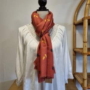 Rostfärgad halsduk med mönster - En rostfärgad halsduk med små mönster i svart och gult. Halsduken har fransar i ändarna och är tillverkad i ett mjukt material.