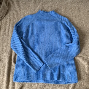 Blå stickad tröja - En bekväm blå stickad tröja med ribbad struktur. Tröjan har långa ärmar och en hög halsringning.