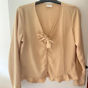 Beige blus med rosett - En beige blus från Vila med en knytning och knappar framtill samt volangdetalj vid nederkanten. Aldrig använd, storlek 38. 🤍✨🌸