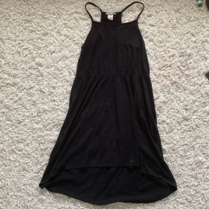 Svart klänning med smala axelband - En svart klänning med smala axelband och en lös passform. Klänningen har en enkel design och är tillverkad i ett mjukt material. Den är knälång och har en något längre bakdel.