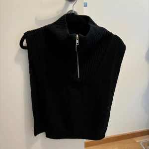 Svart stickad väst med dragkedja - En svart stickad väst med dragkedja framtill. Västen har en hög krage och är ärmlös, vilket gör den perfekt för lager-på-lager under kallare säsonger.