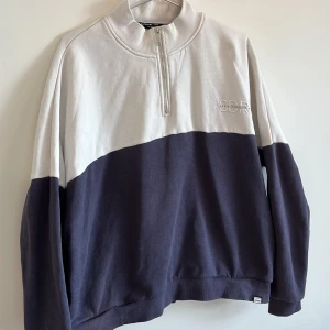 Vit och mörkblå tröja från Superdry - En bekväm vit och mörkblå tröja från Superdry med halv dragkedja vid halsen. Tröjan har långa ärmar och ribbstickade muddar vid ärmslut och nederkant. Märket Superdry är broderat på bröstet. Sitter som M. I använt skick