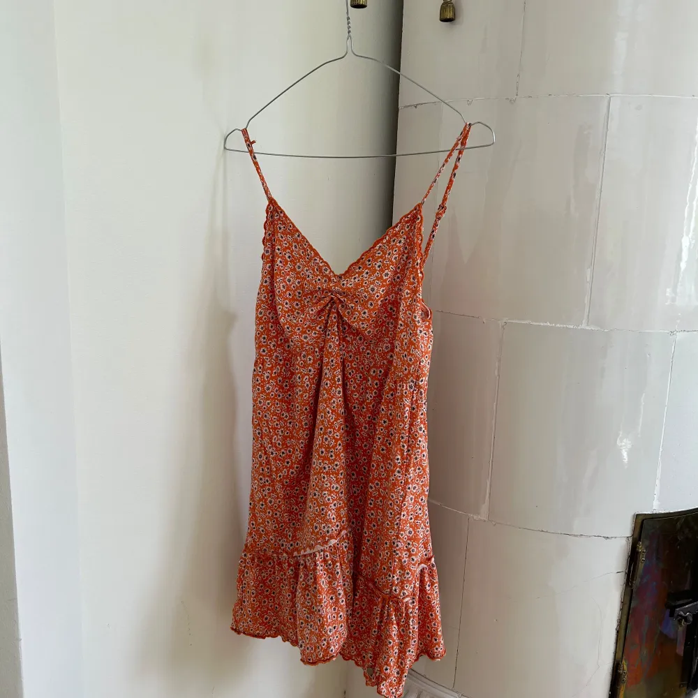 En somrig orange klänning med blommigt mönster. Klänningen har smala axelband och en volangdetalj vid fållen. . Mekot.