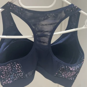 Mörkblå sporttopp med rosa mönster - En mörkblå sporttopp med rosa mönster. Toppen har en racerback-design och är tillverkad i ett stretchigt material som ger bra stöd under träning. aldrig används för att jag har för många.