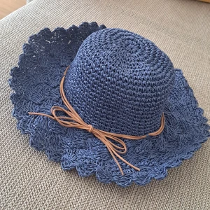 Blå stickad hatt med rosett - En blå stickad hatt med en dekorativ rosett i brunt band. Hatten har en bred, vågig brätte och är tillverkad av ett flätat material.