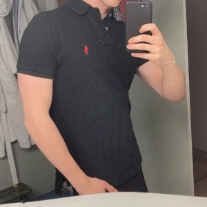 Ralph Lauren piké, Svart - Svart piké från ralph lauren med rött märke, i modell ”custom slim fit”. Köpt i Ralph Lauren på NK för 1300kr. Piken är använd endast ett fåtal gånger och är i bra skick.