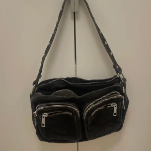 Noelle axelväska i svart mocka - Mocka väska med silver ditaljer. Fint skick! Har också ett långt axelrem som man kan sätta på, om man vill ha den som en axelremsväska, Märke: Noella 