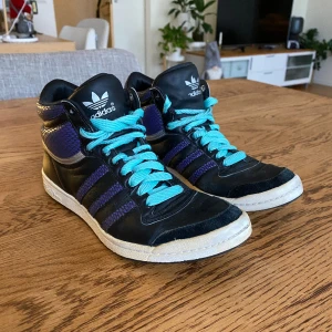 Adidas skor - Svarta adidasskor med lila ränder och turkosa skosnören. Endast använda inomhus så relativt bra skick. Det vita gummit längst ner är aningens gulnat, ena skosnöret har tappat plastpluppen och på vissa ställen är lädret lite slitet, allt syns på bild 3.