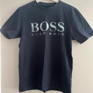 Mörkblå t-shirt från Hugo Boss - En mörkblå t-shirt från Hugo Boss med korta ärmar och en rund halsringning. T-shirten har en stor, glittrig BOSS-logotyp på framsidan.