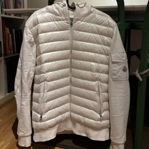 Moncler vit jacka, storlek L - En vit jacka med huva och dragkedja. Jackan har en ficka på vänster ärm och två fickor på framsidan. Jackan är i gott skick.