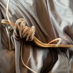 Guldiga högklackade sandaler med snörning - Ett par eleganta guldiga högklackade sandaler med snörning. Skorna har en spetsig tå och en glittrig detalj på snörningen. aldrig använda 