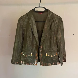 Grön kavaj från Derhy - Derhy- En grön Blazer/Jacket, i boho med dekorativa paljetter längs fållen och fickorna. Kavajen är tunn, är sydd mestadels i linne och. har en glansig yta. -Material: Ytter: 55% Linne, 45%Bomull Inner: 100% Acetat  -Storlek: Medium  Mått- Längd: 60cm Axlar: 40cm Byst: 46cm Midja: 42cm Ärm: 33cm -Skick: Mycket bra