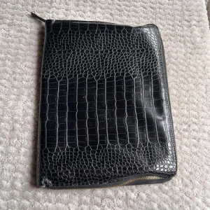 Svart datorfodral med krokodilmönster - En elegant svart datorväska med krokodilmönster. Väskan har en dragkedja längs ena sidan och är tillverkad av låtsasskinn. Perfekt för att bära dokument och dator eller som en stilren accessoar.