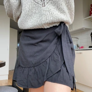 Svart omlottkjol - Svart omlottkjol från Nelly. Knapp på insidan man sätter ihop sidorna med och sen ett rep som man knyter själv. Storlek 38 men tack vare bandet kan man spänna in eller knyta lösare beroende på hur man vill passformen ska vara. Nyskick.