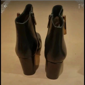 Svarta stövletter från Michael Kors - Eleganta svarta stövletter från Michael Kors med klack och dekorativt spänne på sidan. Skorna har en dragkedja för enkel på- och avtagning.