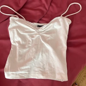 Vit linnetopp - En vit linnetopp med smala axelband och spetsdetaljer vid halsringningen. Toppen har en åtsittande passform och är tillverkad i ett mjukt material.