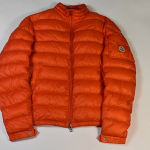 Moncler Lambot Giubbotto - MONCLER LAMBOT GIUBBOTTO - Retail: 6585 SEK - Pris: 2499 SEK - Storlek: 1 (Motsvarar Small) - Färg: Ljus orange - Skick: 8,5/10, finns liten pinhole (Bild 4) Tags ingår  och knapparna har repor - Material: 100% Polyamide, 90% Down, 10% Feather  