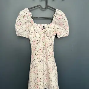 En söt blommig klänning från H&M med puffärmar och en smockad överdel. Klänningen har en vit bas med ett mönster av rosa, lila och gröna blommor. Den är kort och har en tight passform.