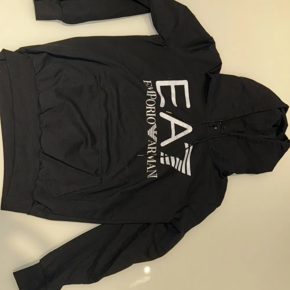 En svart hoodie från Emporio Armani med en stor logotyp på framsidan. Hoodien har en stor ficka på magen och en justerbar huva med dragsko. Den har långa ärmar och ribbstickade muddar vid ärmslut och nederkant.. Tröjor & Koftor.