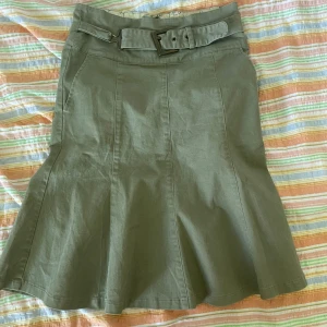 Grön kjol från Vero Moda - En grön kjol från Vero Moda med ett bälte i midjan. Kjolen har en A-linjeformad design och är knälång. Den har en dragkedja baktill för enkel på- och avtagning.