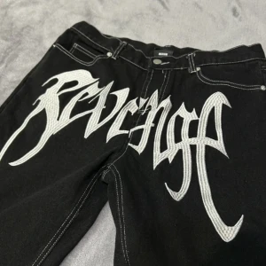 Svarta Revenge Jeans i Storlek Medium - Äkta och Trendiga! - Dessa svarta jeans från det populära märket Revenge är en perfekt kombination av stil och komfort. Tillverkade av högkvalitativt material erbjuder de en perfekt passform och en tidlös estetik. Den klassiska svarta färgen gör dem mångsidiga och lämpli