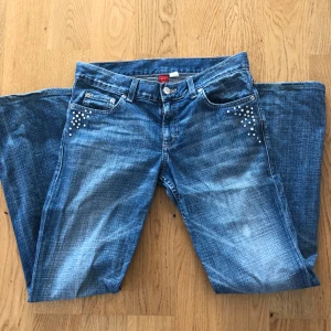Blå jeans med pärldetaljer från Divided - Säljer ett par snygga blå jeans från Divided i storlek 28. De har en låg midja och bootcut-stil, vilket ger en cool och avslappnad look. Jeansen är dekorerade med diamnter på fram- och bakfickorna, vilket ger dem en unik touch. De är tillverkade av 98% bomull och 2% elastan, vilket gör dem både bekväma och stretchiga. Hyfsat skick då några av diamanterna fallit av och en liten blekt fläck på höger lår (se bild, syns nt så mkt) 🩵⭐️