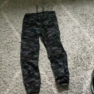 Camouflage mjukisbyxor - Säljer ett par bekväma mjukisbyxor med camouflage-mönster. Byxorna har en dragsko i midjan för justerbar passform och är perfekta för en avslappnad stil. De är i bra skick och redo för nya äventyr!