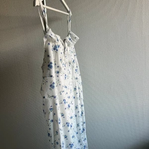 Vit blommig sommarklänning - Säljer en superfin vit sommarklänning med blå blommor. Klänningen har smala axelband och en söt volangdetalj vid bysten. Perfekt för varma sommardagar eller en mysig picknick i parken. Den är i nyskick och har en normal passform. Materialet känns lätt och luftigt, vilket gör den bekväm att bära hela dagen.