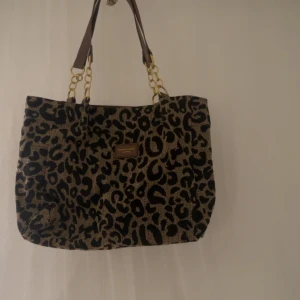 Leopardmönstrad shoppingväska med kedjedetaljer - Säljer en snygg leopardmönstrad shoppingväska med guldfärgade kedjedetaljer på handtagen. Väskan är rymlig och perfekt för shopping eller vardagsbruk. Den har bruna handtag och en liten logotyp på framsidan. Väskan är i bra skick och redo för en ny ägare!