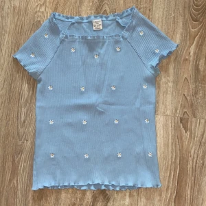Blå ribbad topp från Lindex - Säljer en söt blå ribbad topp från Lindex i storlek 146/152. Toppen har korta ärmar och små vita blommor broderade över hela tyget. Den har en fin volangkant vid ärmslut och nederkant. Perfekt för sommarens varma dagar! 🌸