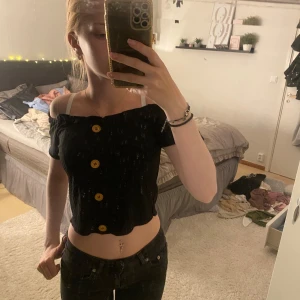 Svart off-shoulder topp med knappar - Säljer en snygg svart off-shoulder topp med stora gula/guldiga knappar framtill. Toppen är kortärmad och har en tight passform. Perfekt för sommarens fester eller en utekväll med vänner. Toppen är i bra skick och redo för en ny ägare!