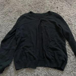 Svart kofta från H&M - En svart kofta från H&M med knappar framtill. Koftan har långa ärmar och ribbstickade muddar vid ärmslut och nederkant.