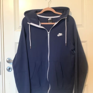 Mörkblå hoodie från Nike - Säljer en snygg mörkblå hoodie från Nike i storlek S. Den har en dragkedja framtill och en vit Nike-logga på bröstet. Hoodien har en justerbar huva med vita snören och är tillverkad i ett mjukt och bekvämt material. Perfekt för både träning och vardagsbruk!