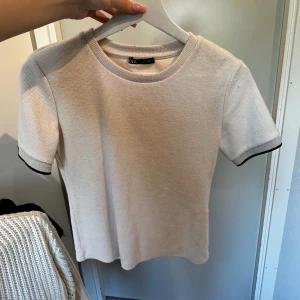 Vit kortärmad topp från Zara - En vit kortärmad topp från Zara med svarta kanter vid ärmslut och halsringning. Toppen har en ribbad struktur och är tillverkad i ett bekvämt material.