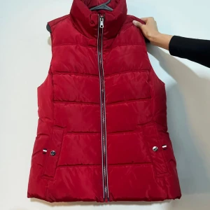 Röd dunväst från Tommy Hilfiger - En röd dunväst från Tommy Hilfiger med dragkedja framtill och två fickor med knappar. Västen har en hög krage och är i storlek S.