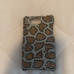 Glittrigt mobilskal med leopardmönster - Ett glittrigt mobilskal dekorerat med ett leopardmönster i silver, svart och orange. Skalet är täckt med små pärlor som ger en glittrande effekt.