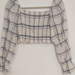 Rutig blus med långa ärmar - En rutig blus med långa ärmar. Blusen har en elastisk, rynkad design på kroppen med ihopdragningar på ärmarna. croptop. Färgerna är vit och blå rutigt