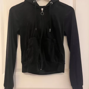 Svart hoodie med dragkedja - En svart hoodie med dragkedja framtill. Jackan har en huva med dragsko och långa ärmar. Materialet ser ut att vara mjukt och bekvämt.