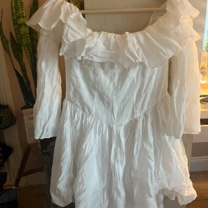 Vit klänning med volanger - En vit klänning med volanger och långa ärmar. Klänningen har en offshoulder-design och är tillverkad i ett lätt och luftigt material.