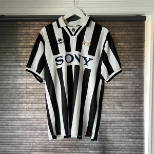 Svartvit randig fotbollströja från Kappa - En svartvit randig fotbollströja från Kappa med korta ärmar. Tröjan har en krage och två stjärnor ovanför logotypen 'SONY' på framsidan. Kappa-logotypen är broderad på bröstet.