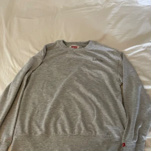 Grå tröja från Levi's - En grå långärmad tröja från Levi's med rund halsringning. Tröjan har en enkel design med Levi's logotyp broderad på bröstet och en liten röd etikett på ärmen.