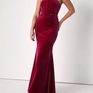 Långklänning röd/rosa Strl L - Klänning med korsett från Lulus i mörkröd sammet, lite åt det rosa hållet. Super stretchigt framförallt över mage och höfter, även korsetten sitter väldigt bekvämt. Faller fint tack vare det tunga tyget. Tycker personligen att den kan upplevas lite stor i storleken pga stretchen. Går även att använda som gravid. Perfekt för gäster på vinterbröllop! Använd endast en gång och i super bra skick. 