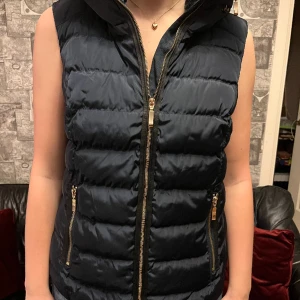 Väst  - Varm väst, aldrig använd  Färg mörkt blå med guldfärgade dragkedjor. Luva finns dold i kragen. Storlek 40  Prisförslag  Frakt tillkommer 
