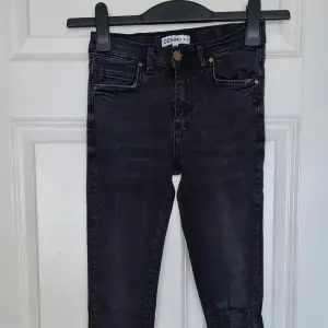 Ett par svarta jeansbyxor från märket Denim by Skill. Byxorna har en tight passform och är dekorerade med en reva på ena benet. De har en knapp och dragkedja framtill samt fem fickor. Byxorna har en rå kant vid bensluten.