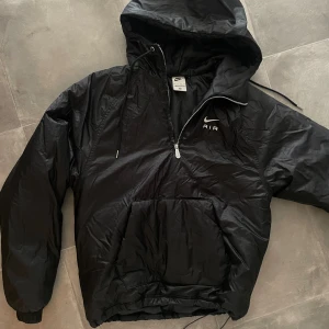 Nike jacka - Det här är en svart Nike Air-jacka med huva. Jackan är en halvlång modell med en dragkedja som går upp till bröstet. Den har en stor ficka framtill, som också fungerar som handvärmare. Jackan har dragskor i nederkanten för att justera passformen, 