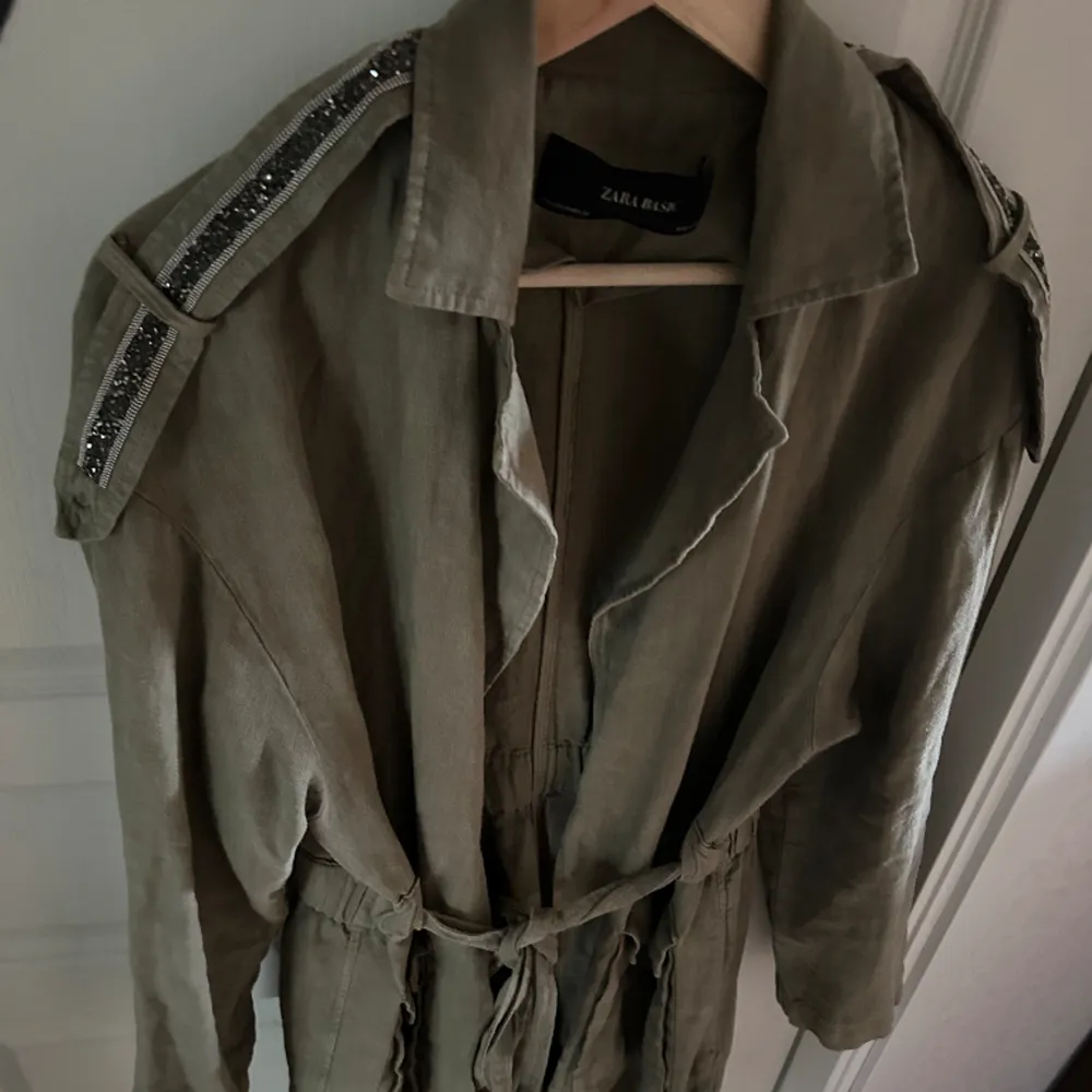 Khaki jacka i 100% linne med detaljer av stenar. Knyts i midjan och går till mitten av låret ungefär. . Takit.