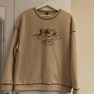 Tröja i beige med bruna sömmar  - Beige tröja med brunna sömmar och ängeltryck 